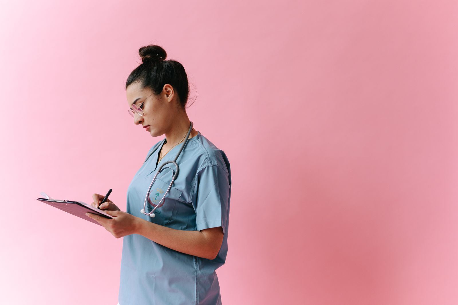 A clinician in scrubs with a stethoscope writing notes on a clipboard